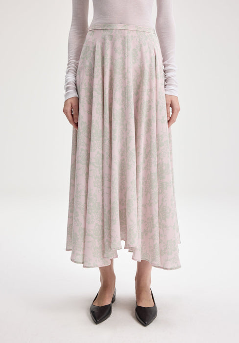 Close-up of a flowy, mid-length pink skirt with a soft green floral pattern, worn with a sheer white long-sleeve top and black pointed-toe flats.