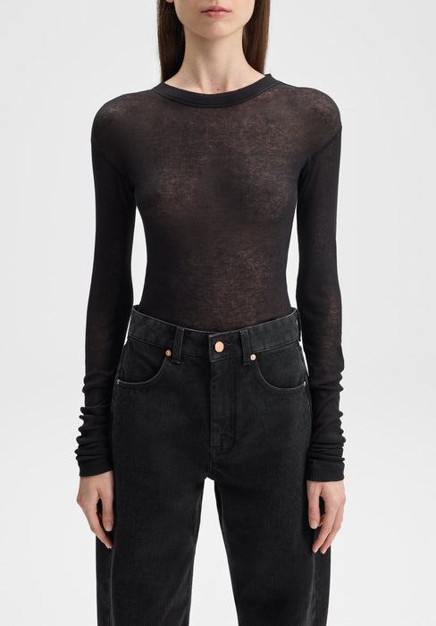 Front-facing close-up of a model from neck to hips, standing upright with arms relaxed at her sides, wearing a sheer black long-sleeve top paired with high-waisted black jeans against a plain background.