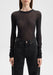 Front-facing close-up of a model from neck to hips, standing upright with arms relaxed at her sides, wearing a sheer black long-sleeve top paired with high-waisted black jeans against a plain background.