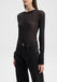 Close-up of a model from shoulders to hips wearing a sheer black long-sleeve top over dark high-waisted jeans, standing slightly angled with one arm relaxed by her side against a plain background.