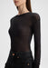 Close-up side view of a model from shoulders to hips, slightly angled to the left, wearing a sheer black long-sleeve top tucked into high-waisted black jeans, with one arm relaxed along her side against a plain background.