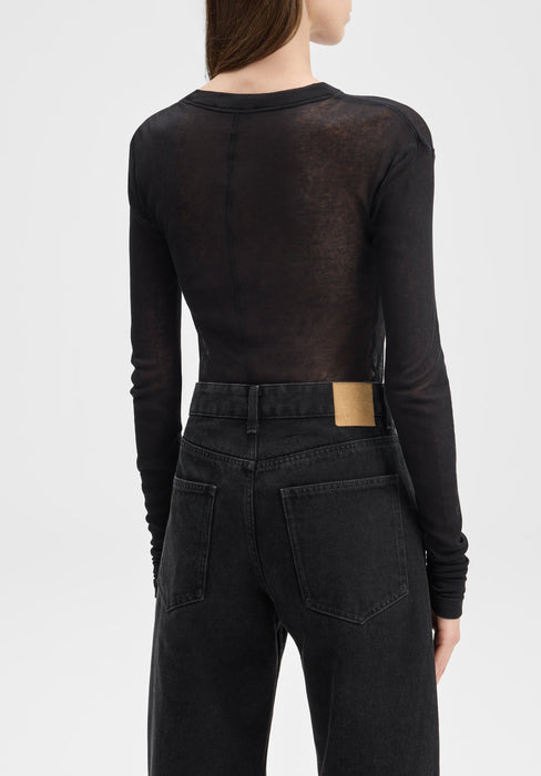 Back view of a model from shoulders to hips, standing upright with arms relaxed at her sides, wearing a sheer black long-sleeve top tucked into high-waisted black jeans against a plain background.