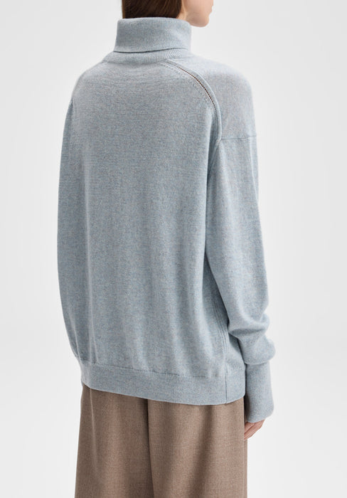 Back-facing view of the model standing upright with her body slightly angled to the side, arms relaxed at her sides, showcasing the rear of a light blue oversized turtleneck sweater paired with beige trousers.