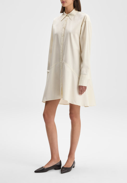 Full-length three-quarter view of a model standing with one leg slightly forward, wearing a cream long-sleeve button-down shirt dress with a flared hem and pointed collar, paired with black flats against a plain background.