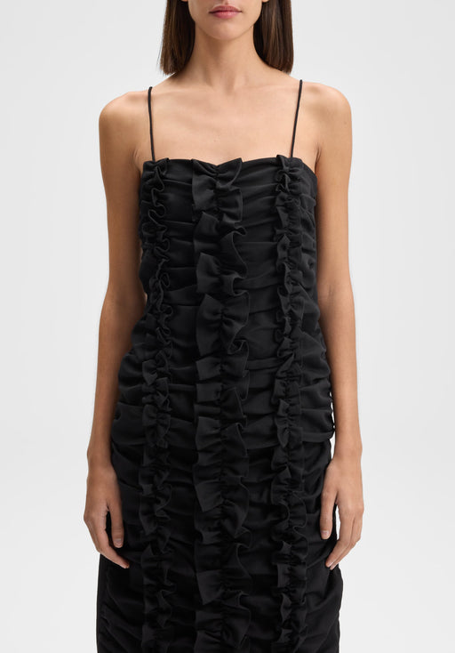 Front-facing view of the model standing upright with shoulders squared and arms relaxed straight at her sides, wearing a black spaghetti-strap dress with vertical ruffle detailing down the front.