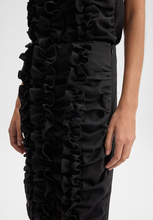 Close-up detail of a black dress showing vertical ruffle embellishments along the fabric, with the model’s hands relaxed at her sides against a plain background.