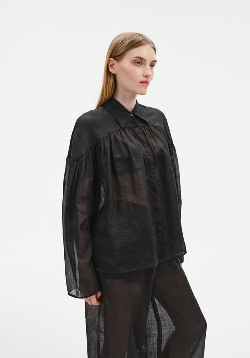Three-quarter side view of a model wearing a sheer black long-sleeve blouse with a pointed collar and gathered yoke, paired with matching translucent trousers, highlighting the airy fabric and relaxed fit.