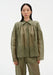 Front-facing view of a model standing upright with arms relaxed at her sides, wearing a sheer olive-green blouse with a pointed collar and gathered detailing, paired with matching lightweight trousers, emphasizing the soft, airy fabric and straight silhouette.