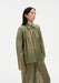 Three-quarter side view of a model standing with a slight lean and one leg forward, wearing a sheer olive-green blouse with a pointed collar and gathered yoke, paired with matching lightweight trousers, highlighting the soft drape and airy fabric.