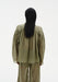 Back-facing view of a model standing upright with arms relaxed at her sides, wearing a sheer olive-green blouse with gathered yoke and long sleeves, paired with matching translucent trousers, showing the loose drape from behind.