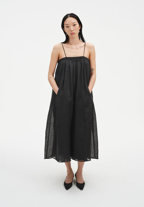 A woman stands facing forward with a relaxed posture against a plain white background, wearing a black sleeveless midi dress with thin straps and a loose, gathered silhouette. Her hands are in her pockets, her dark shoulder-length hair is worn down, and she tilts her head slightly with a neutral expression, paired with black pointed heels.