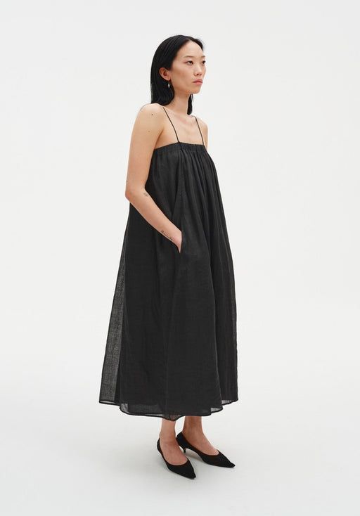 A woman stands in a relaxed three-quarter side pose against a plain white background, wearing a black sleeveless midi dress with thin straps and a loose, gathered silhouette. She has one hand in her pocket, her dark shoulder-length hair worn down, and she looks slightly to the side with a neutral expression, paired with black pointed heels.