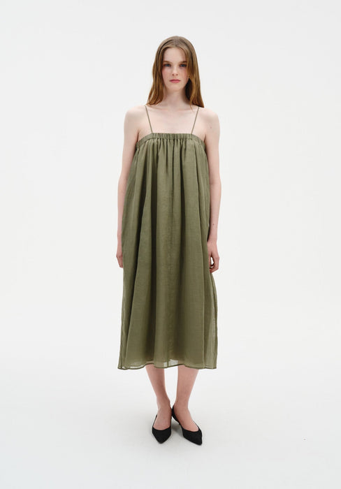 A woman stands facing forward in a straight, centered stance with feet close together, wearing an olive-green sleeveless midi dress with thin straps and a loose, gathered silhouette, paired with black pointed heels.