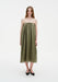 A woman stands facing forward in a straight, centered stance with feet close together, wearing an olive-green sleeveless midi dress with thin straps and a loose, gathered silhouette, paired with black pointed heels.