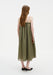 Rear view of a woman standing with a slight turn to one side, her weight subtly shifted, wearing an olive-green sleeveless midi dress with thin straps and a loose, gathered silhouette.