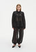 A woman stands facing forward against a plain white background, wearing a sheer black long-sleeve blouse with a collar and gathered detailing, paired with matching semi-transparent billowy trousers and black pointed heels.