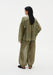 Rear three-quarter view of a woman against a plain white background, wearing a loose olive-green sheer blouse with gathered detailing across the back and matching lightweight trousers with a relaxed, billowy fit.