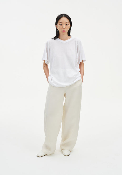 A woman stands facing forward in a relaxed stance against a plain white background, wearing an oversized white T-shirt and loose cream-colored wide-leg trousers with light-colored shoes, her hands in her pockets and posture slightly casual.