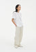 A woman stands in a relaxed three-quarter side pose against a plain white background, wearing an oversized white T-shirt tucked into loose cream-colored trousers and light-colored shoes. Her dark shoulder-length hair is worn down, and she looks slightly off to the side with her hands in her pockets.
