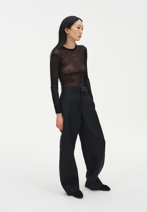 A woman stands in a relaxed three-quarter side pose against a plain white background, wearing a sheer long-sleeve dark top and black wide-leg trousers with black shoes. Her dark shoulder-length hair is worn down, and she looks slightly to the side with a neutral expression, one hand resting in her pocket.