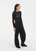 A woman stands in a relaxed three-quarter side pose against a plain white background, wearing a sheer long-sleeve dark top and black wide-leg trousers with black shoes. Her dark shoulder-length hair is worn down, and she looks slightly to the side with a neutral expression, one hand resting in her pocket.