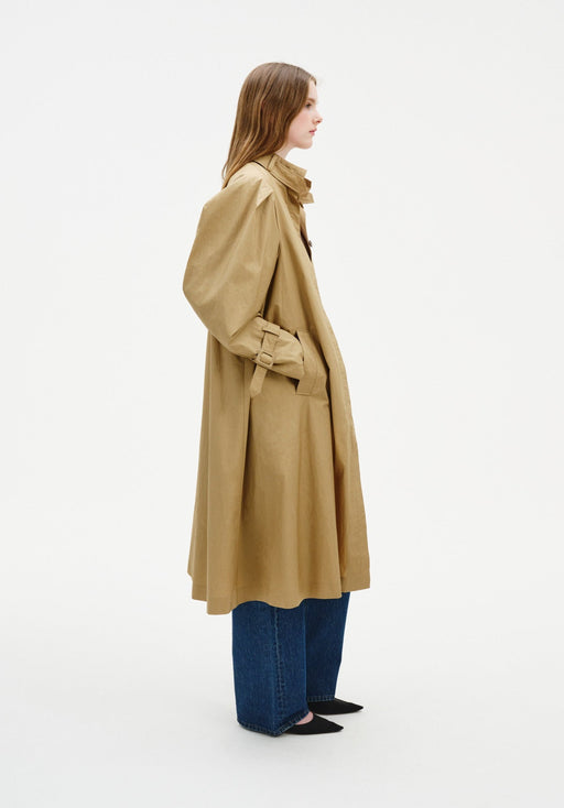 Side view of a model wearing a beige trench-style coat with voluminous sleeves and cuff straps, layered over blue jeans and black pointed shoes.