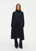 Front view of a model wearing a long, black, semi-sheer coat-style dress with a high neckline, long sleeves, and large patch pockets, layered over matching wide-leg trousers and paired with black pointed shoes.