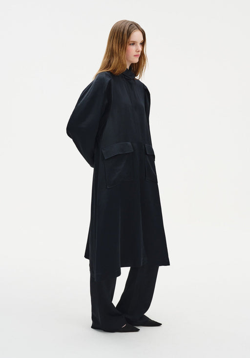 Side view of a model wearing a long black coat-style dress with a high neckline, voluminous sleeves, and large front patch pockets, layered over matching wide-leg trousers and paired with pointed black shoes.