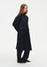 Side view of a model wearing a long black coat-style dress with a high neckline, voluminous sleeves, and large front patch pockets, layered over matching wide-leg trousers and paired with pointed black shoes.