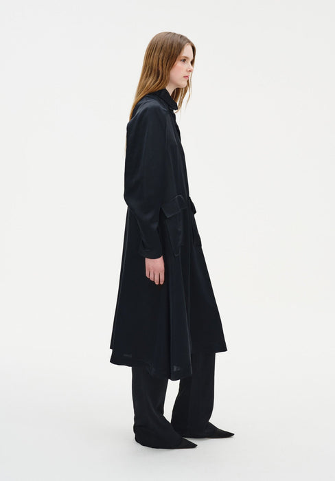 Side profile of a model wearing a long black coat-style dress with a structured collar, relaxed sleeves, and a loose, flowing silhouette, layered over matching wide-leg trousers and paired with pointed black shoes.