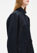Close-up side view of a black coat-style dress featuring a structured stand collar, subtle sheen fabric, voluminous sleeves, and a large front patch pocket detail.