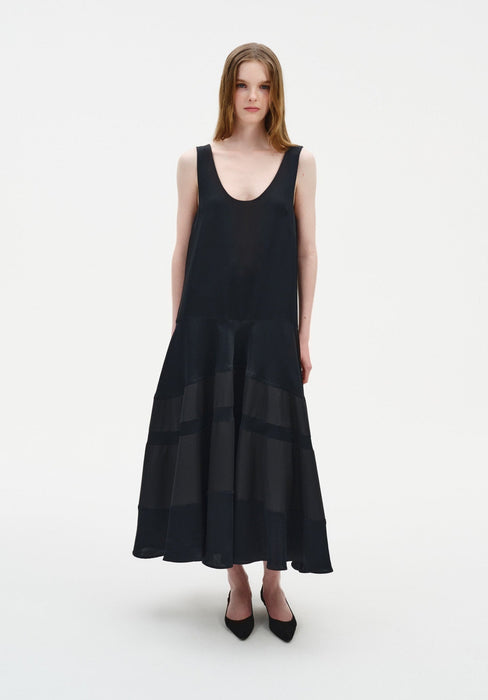Front view of a model wearing a sleeveless black midi dress with a scoop neckline and a softly flared, semi-sheer panelled skirt, paired with black pointed shoes.
