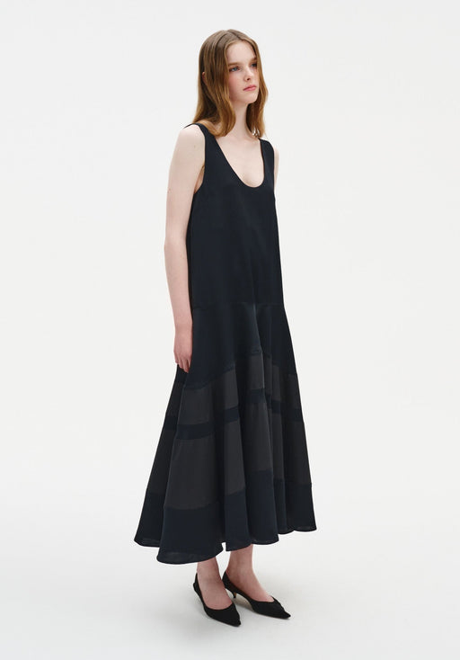Side view of a model wearing a sleeveless black midi dress with a scoop neckline and a softly flared, semi-sheer panelled skirt, paired with black pointed shoes.