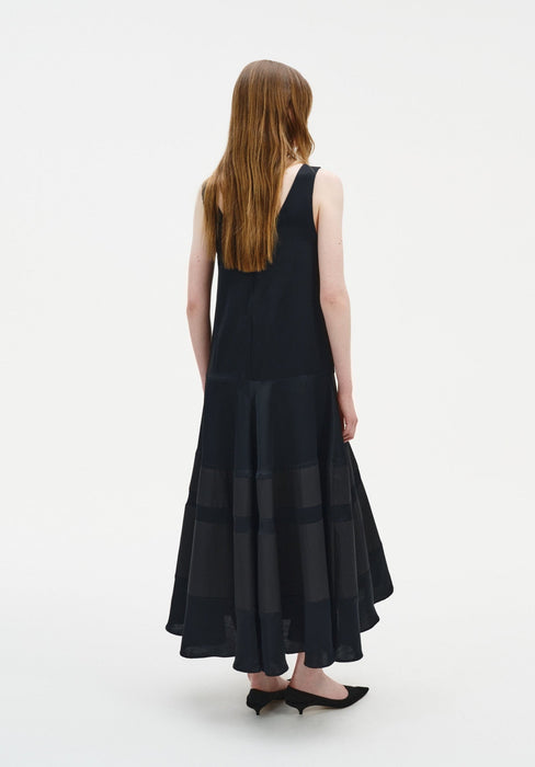 Back view of a model wearing a sleeveless black midi dress with a scoop back and a softly flared, semi-sheer panelled skirt, paired with black pointed shoes.