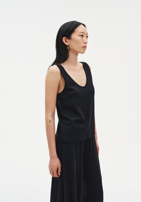 Side view of a woman standing in a relaxed posture, wearing a black sleeveless tank top with a scoop neckline and matching loose-fitting trousers, set against a plain studio background.