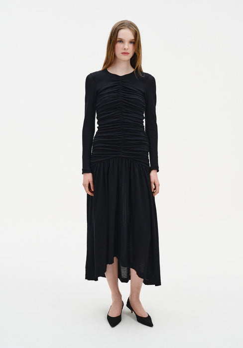 A woman stands facing forward against a plain white background, wearing a black long-sleeve dress with ruched detailing on the bodice and a loose, mid-calf skirt. She has straight, light brown hair worn down and is dressed with black pointed-toe heels, maintaining a neutral expression with arms relaxed at her sides.