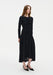 A young woman with long light brown hair stands against a plain white background, wearing a fitted black long-sleeve dress with ruched detailing and a flowy mid-calf skirt, paired with black pointed heels. She faces slightly to the side with a neutral expression and relaxed posture.