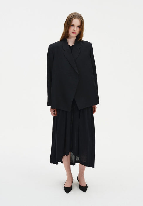 A woman stands against a plain white background wearing an oversized black blazer layered over a black ruched dress with a mid-calf skirt, paired with black pointed heels. She faces forward with a slight tilt of the head and a neutral expression, her straight light brown hair worn down.