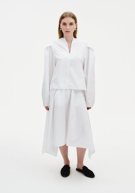 Front view of a woman wearing a crisp white long-sleeve top with a notched neckline and structured shoulders, paired with a white asymmetrical midi skirt and black slip-on flats.
