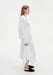 Side view of a woman wearing a white long-sleeve top with a clean, minimal silhouette paired with a white asymmetrical midi skirt, styled with black slip-on flats.