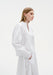 Side view of a model wearing a white blouse with a V-slit neckline and voluminous sleeves, paired with a matching flowing skirt, hands in pockets.