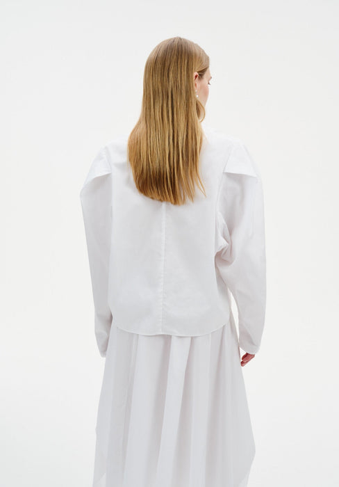 Back view of a model wearing a white blouse with structured shoulders and long sleeves, paired with a flowing white skirt, shown against a plain background.
