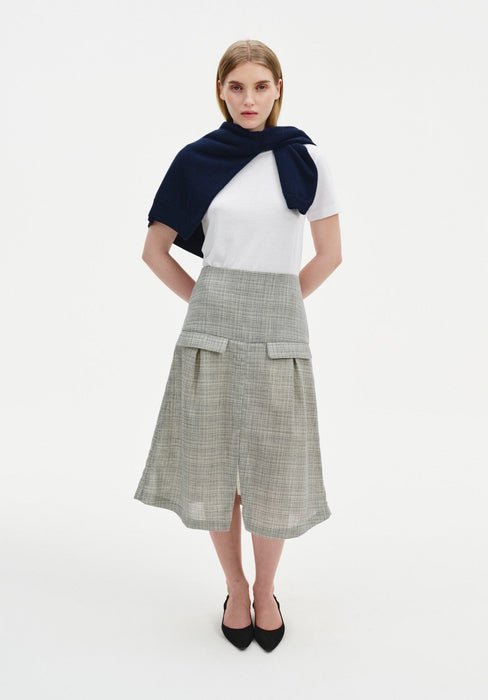 A woman stands facing forward with a slight three-quarter angle against a plain white background, wearing a white short-sleeve top with a navy sweater draped over her shoulders, paired with a light gray textured midi skirt with flap pockets and a front slit, and black pointed heels.
