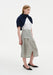 A woman stands in a three-quarter side pose against a plain white background, wearing a white top with a navy sweater draped over her shoulders, paired with a light gray textured midi skirt featuring a front slit and flap pocket detail, and black pointed heels. Her straight light brown hair is worn down, and she looks slightly to the side with a neutral expression.