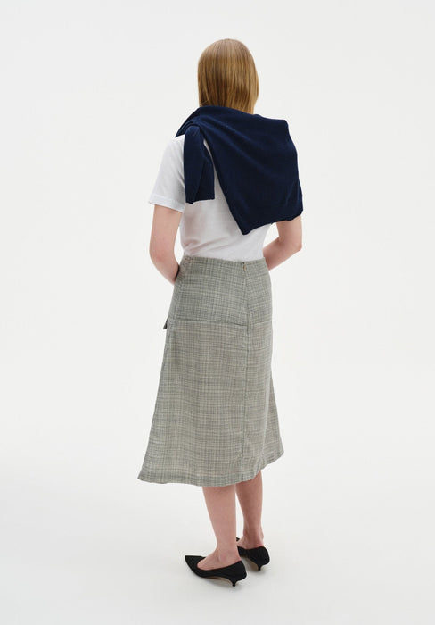 Rear three-quarter view of a woman against a plain white background, wearing a white short-sleeve top with a navy sweater draped over her shoulders, paired with a light gray textured midi skirt and black pointed heels.