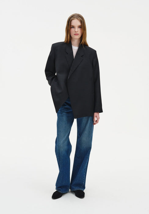 A woman with long light brown hair stands facing forward against a plain background, posing with a relaxed stance, one hand in her pocket, and a neutral expression.