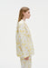 Side profile of a woman wearing a white long-sleeve shirt and matching trousers with a soft yellow floral pattern, highlighting the relaxed fit and clean silhouette against a plain studio background.