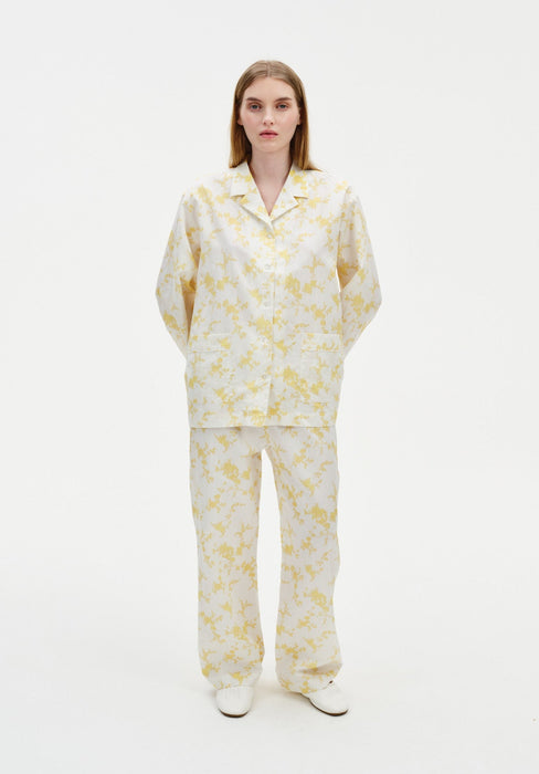 Front view of a model wearing a cream two-piece set with a yellow abstract floral print, featuring a long-sleeve collared button-up top with patch pockets and relaxed straight-leg trousers, paired with white shoes.