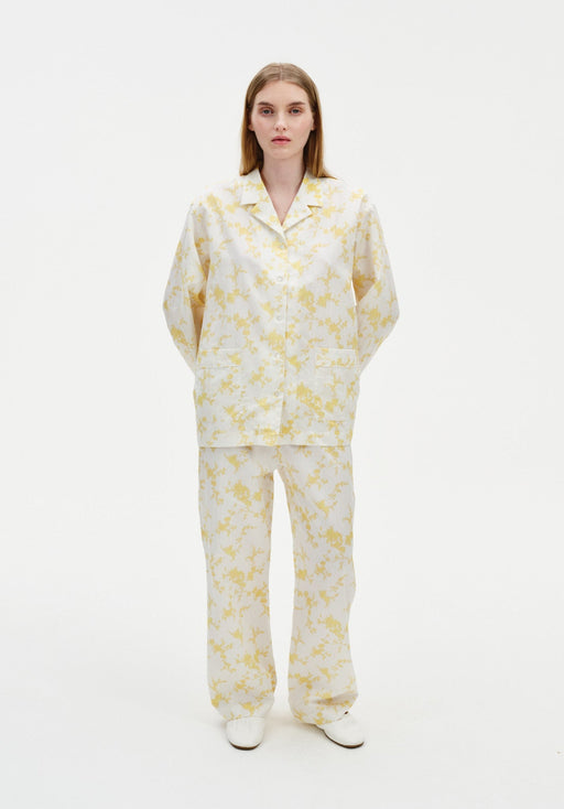 Front view of a model wearing a cream two-piece set with a yellow abstract floral print, featuring a long-sleeve collared button-up top with patch pockets and relaxed straight-leg trousers, paired with white shoes.