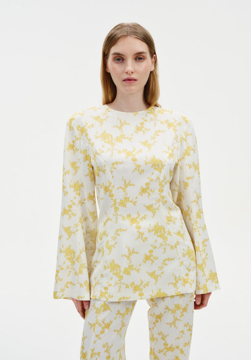 A woman stands facing forward against a plain white background, wearing a cream-colored long-sleeve top with a yellow floral pattern and matching trousers. The top has a relaxed fit with slightly flared sleeves, and she maintains a neutral expression.